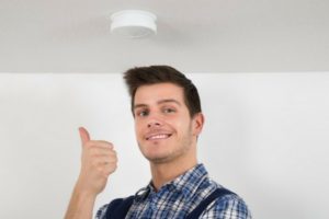 Man giving a thumbs up near a smoke detector, emphasizing home safety and maintenance checks during winter.