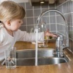 A young child at a kitchen sink reaching for a faucet, with water flowing, illustrating domestic plumbing situations.