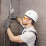 Male plumber installing showerhead and plumbing fixtures in a bathroom with modern tile wall, wearing safety gear.