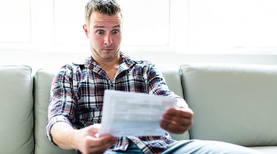 Surprised man reading plumbing-related document on paper, seated on light couch in natural light.