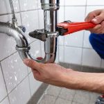 Human hand repairing a leaking plumbing pipe with an adjustable wrench at a tiled floor and wall, by Green Hills Plumbing.