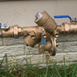 Brass water valve assembly with blue handles near a building foundation, vital for safe plumbing systems.